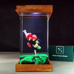 May include: A resin art piece with two anime-style figures inside a clear rectangular block. One figure is in a red dress, the other in a yellow shirt. The base and top are made of wood, with a green moss-like base. The piece is illuminated.