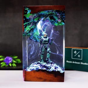 May include: A resin art piece featuring a green action figure of a soldier holding a weapon, set against a backdrop of swirling blue and green colors. The figure stands on a textured base within a clear resin block with a wooden frame.