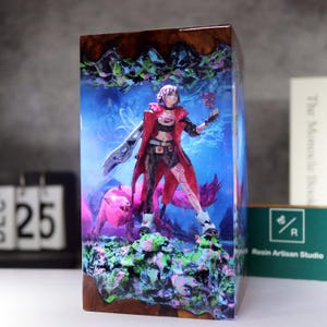 May include: A resin art piece featuring a female figure in a red coat, holding a mask and a sword, standing with a pink wolf. The artwork is encased in a rectangular block with a wooden base and a blue and green background.
