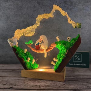 May include: A resin sculpture of a white wolf and a small human figure on a tree branch, surrounded by small white figures with glowing eyes. The sculpture is set in a clear resin base with green moss and a light inside. The base is made of wood and has the text "Resin Artisan Studio" on it.