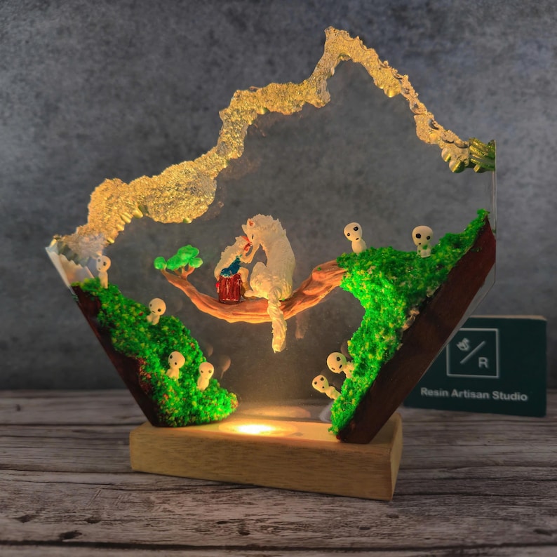 May include: A resin sculpture of a white wolf and a small human figure on a tree branch, surrounded by small white figures with glowing eyes. The sculpture is set in a clear resin base with green moss and a light inside. The base is made of wood and has the text "Resin Artisan Studio" on it.