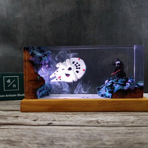 Könnte beinhalten: Ein weißes Millennium-Falken-Raumschiff und eine schwarze Darth-Vader-Figur sind in einem klaren Harzblock mit einem blau-violetten galaktischen Hintergrund eingeschlossen. Der Block wird von unten beleuchtet, wodurch ein leuchtender Effekt entsteht.