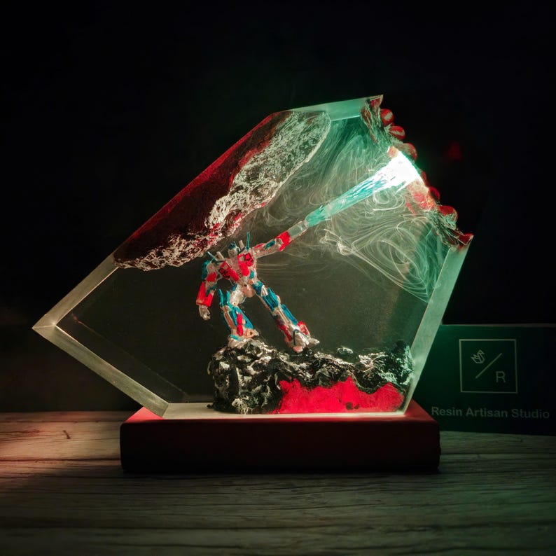May include: A red, blue, and silver robot figurine is encased in clear resin with red and white swirls. The resin is shaped like a diamond and has a green light shining from the robot's hand. The base is red and the text "Resin Artisan Studio" is visible on a green label.