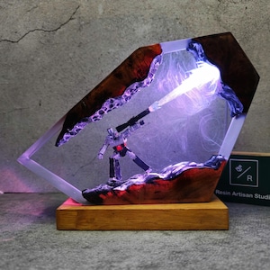 Op de afbeelding: Een harslamp met een robotfiguur in een helder, geometrisch frame. De lamp heeft een houten voet en bevat hout- en paarse accenten. De robot vuurt een lichtstraal af. Decoratief item.