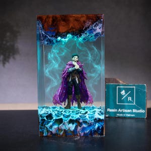 May include: A rectangular resin block containing a miniature figure of a bearded character wearing a purple cloak. The base and top feature textured blue and brown waves, while the centre is filled with swirling translucent teal smoke effects.