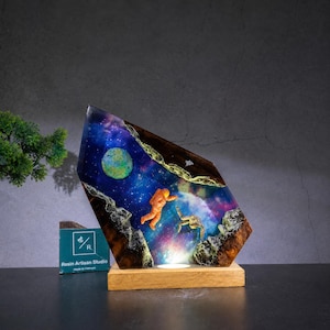 May include: A decorative resin lamp featuring a space scene with an orange astronaut floating in a nebula of blue, purple, and pink colours, with a green planet and dark wood edges, mounted on a rectangular wooden base with a small logo sign.
