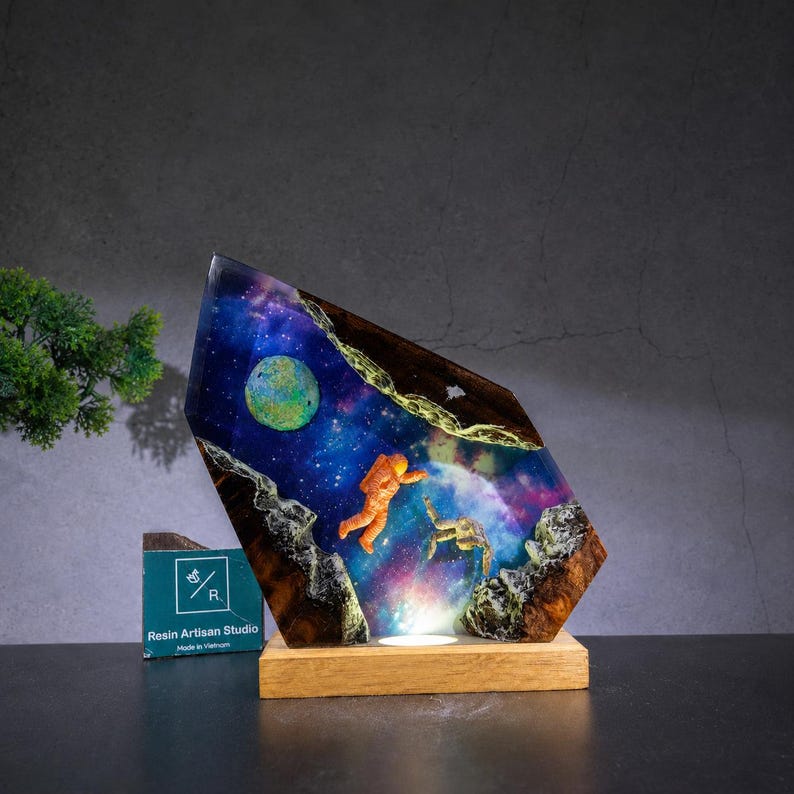 May include: A decorative resin lamp featuring a space scene with an orange astronaut floating in a nebula of blue, purple, and pink colours, with a green planet and dark wood edges, mounted on a rectangular wooden base with a small logo sign.