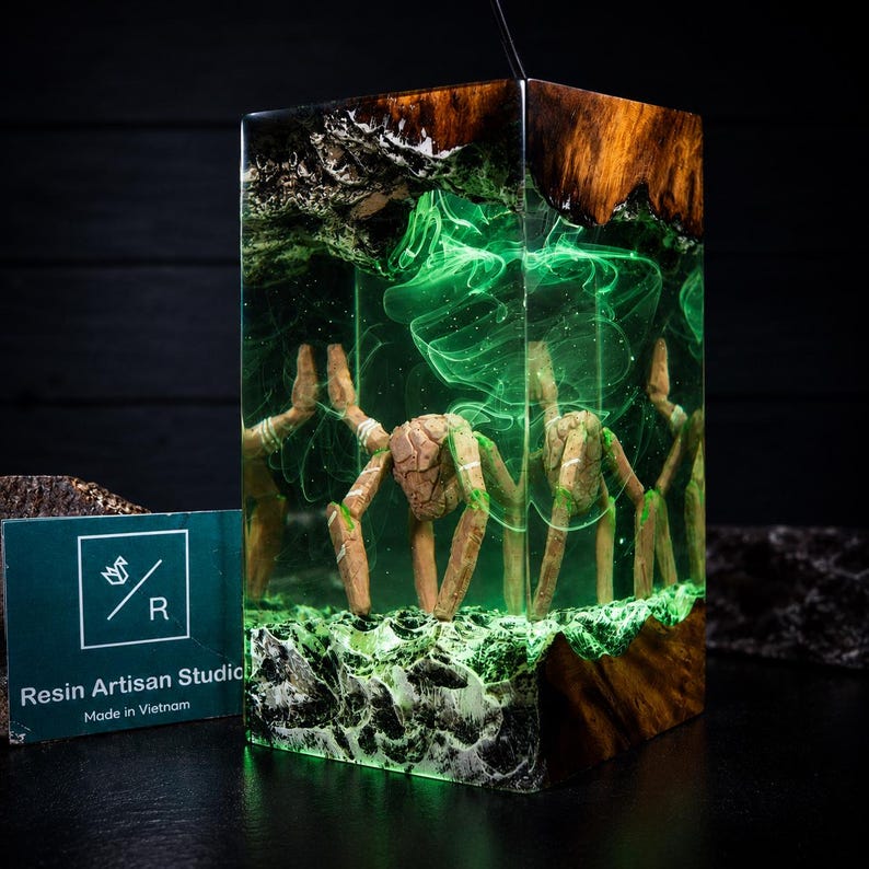 May include: A rectangular resin lamp featuring a wooden base and top with a central, multi-legged creature sculpture suspended in clear resin. The interior glows with a vibrant green light, creating swirling patterns around the figure.
