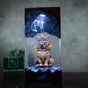 May include: A resin art piece featuring a detailed cat figurine wearing goggles and a crown, set within a clear block. The cat is multi-colored with shades of orange, yellow, and purple. The background has a cave-like design with blue and purple lighting.
