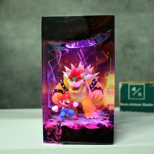 May include: A rectangular resin art piece featuring the characters Mario and Bowser from the Super Mario video game series. Bowser is a large, orange turtle-like creature with spikes, and Mario is in his classic red and blue outfit. The background has a purple and orange glow.