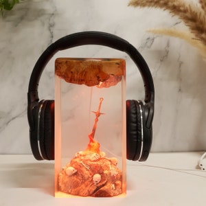 May include: A rectangular resin art piece with a sword on a fiery base with skulls and skeletons. The top section has a wooden texture. The piece glows orange, and black headphones are placed behind it.