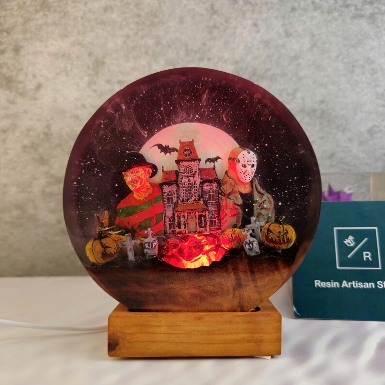 K&ouml;nnte beinhalten: Eine runde, transparente Harzlampe mit Halloween-Motiv. Die Lampe zeigt ein Spukhaus, einen Vollmond, Flederm&auml;use und drei Horrorfilmfiguren: Freddy Krueger, Jason Voorhees und Michael Myers. Die Lampe wird von unten beleuchtet und erzeugt ein warmes Leuchten.