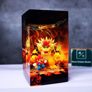 May include: A rectangular resin block featuring a vibrant scene with the characters Bowser and Mario from the Super Mario video game series. The background depicts a fiery, hellish landscape. The block is on a white surface.