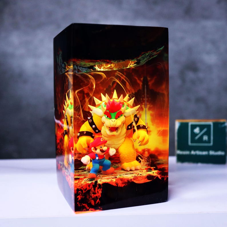 May include: A rectangular resin block featuring a vibrant scene with the characters Bowser and Mario from the Super Mario video game series. The background depicts a fiery, hellish landscape. The block is on a white surface.