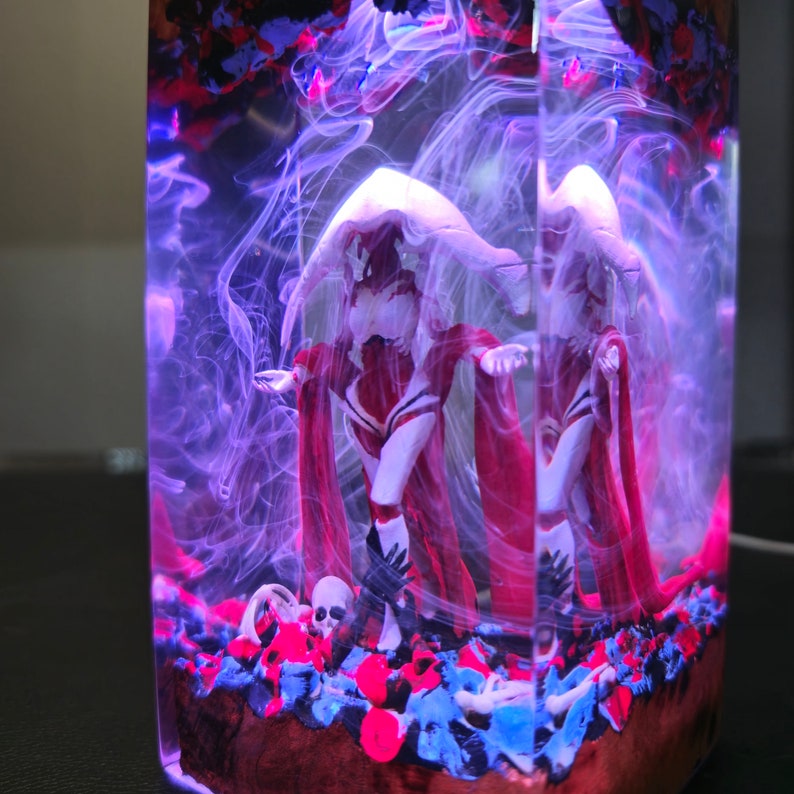 May include: Two white figures with red cloaks are encased in a clear resin block. The figures are standing on a bed of blue, red, and white stones. The resin block is illuminated with purple light.