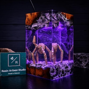 May include: A rectangular resin and wood lamp featuring a glowing purple interior with swirling smoke-like patterns and a central multi-legged creature figure. The base and top are made of dark wood, and a small card reads Resin Artisan Studio Made in Vietnam.