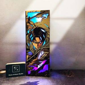 Op de afbeelding: Een rechthoekig, verlicht harskunstwerk met een personage uit de anime-serie Attack on Titan. Het kunstwerk toont een personage met blauwe ogen en een vastberaden uitdrukking, met blauwe en paarse LED-verlichting. Een kleine zwarte doos met tekst staat op de voorgrond.
