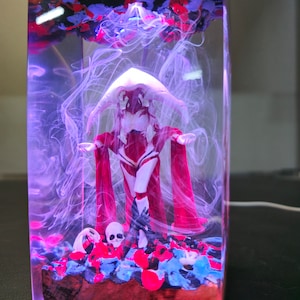 May include: A resin sculpture of a female figure in a red and white dress with a hood. The figure is standing in a clear resin block with pink and blue swirls. The figure is surrounded by small, pink and blue resin flowers.