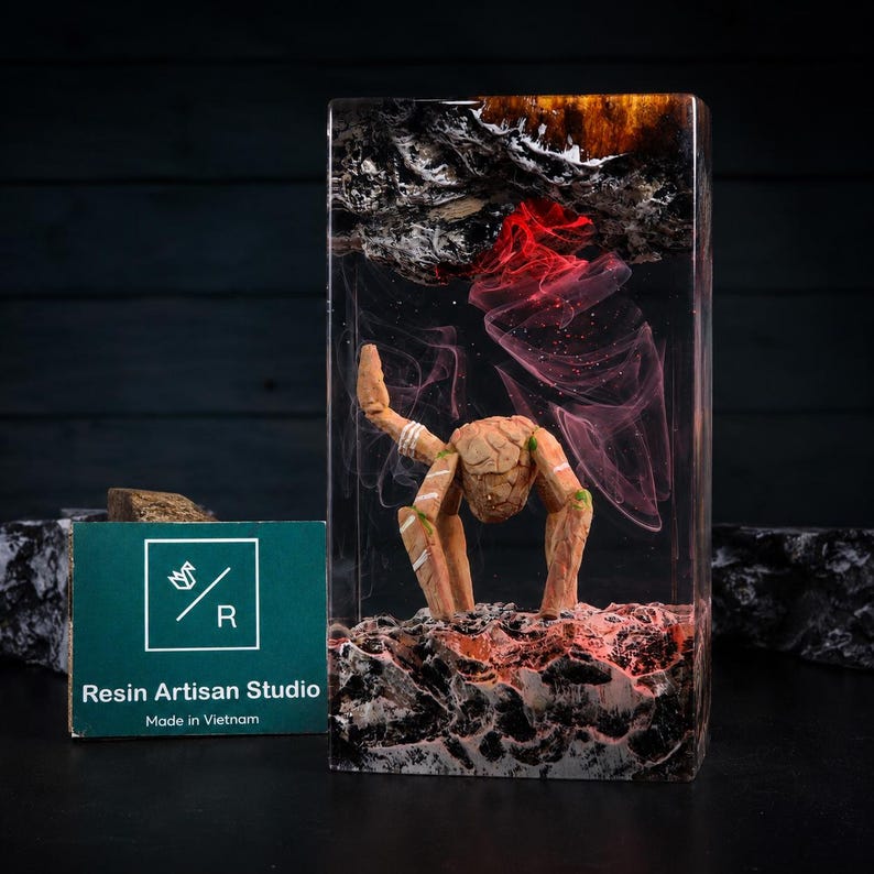 May include: A rectangular resin block featuring a suspended wooden creature with four legs and a textured body. The base and top contain dark, rocky textures with red and white swirling resin effects. A green card displays the text Resin Artisan Studio.