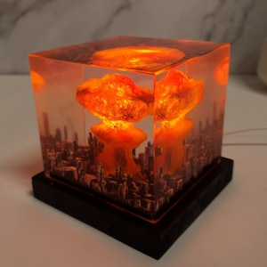 Puede incluir: Una lámpara de cubo transparente con una base negra, que representa una nube en forma de hongo sobre el horizonte de una ciudad. La lámpara está iluminada desde el interior, proyectando un brillo naranja.