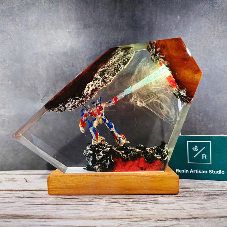 May include: A resin sculpture of a robot holding a glowing blue weapon, encased in a clear diamond-shaped block with a brown and red base. The sculpture is on a wooden base with the text "Resin Artisan Studio" on a green card.