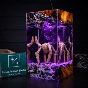 May include: A rectangular lamp made of wood and purple resin, featuring a central figure with multiple limbs resembling a spider or creature. The resin contains swirling purple smoke effects and light, with a dark wood top and base.