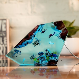 May include: A blue resin sculpture of an underwater scene with sea turtles, an octopus, and divers. The sculpture is shaped like a diamond and has a wood-like texture on the top.