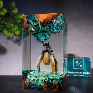 May include: A rectangular resin and wood block sculpture featuring a miniature dark figure riding a tan, multi-legged creature. The piece includes swirling teal and green resin accents resembling water or terrain, with a Resin Artisan Studio logo tag.