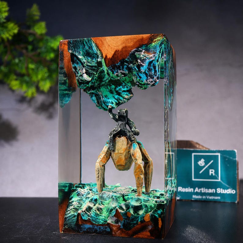 May include: A rectangular resin and wood block sculpture featuring a miniature dark figure riding a tan, multi-legged creature. The piece includes swirling teal and green resin accents resembling water or terrain, with a Resin Artisan Studio logo tag.