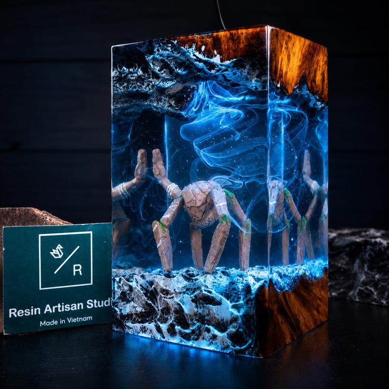May include: A rectangular resin lamp featuring a wooden top and base with a central scene of a multi-legged creature submerged in swirling blue light. A green card with the text Resin Artisan Studio and a white origami logo is placed to the left.