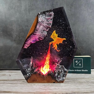May include: A resin art piece with a dragon breathing fire in a starry night sky. The piece is lit from within, creating a warm glow. The piece is made by Resin Artisan Studio.