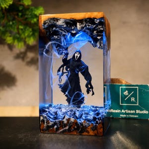 Könnte beinhalten: Eine rechteckige Lampe aus Harz und Holz mit einer Ghostface-Figur, die in klarem Harz mit blauen, wirbelnden Raucheffekten eingeschlossen ist. Basis und Oberseite bestehen aus Naturholz, inklusive einer grünen Karte von Resin Artisan Studio.