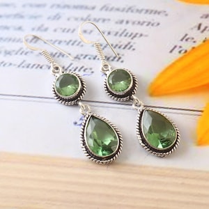 Op de afbeelding: Een paar zilveren oorhangers met groene edelsteen druppels. De oorbellen hebben een traanvorm en zijn versierd met een kleinere ronde edelsteen.