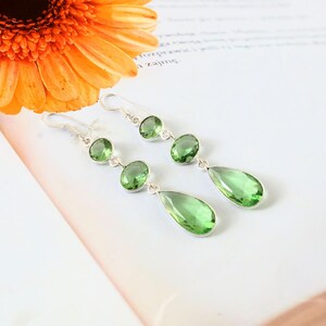 Op de afbeelding: Een paar zilveren oorbellen met groene edelstenen. Elke oorbel heeft drie ronde stenen en een druppelvormige steen. De oorbellen worden getoond op een wit oppervlak, met een oranje bloem op de achtergrond.