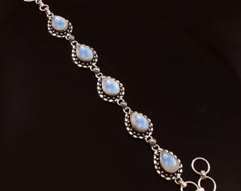 Op de afbeelding: Zilveren armband met vijf peervormige, iriserende maansteen cabochons. Elke steen is gezet in een decoratief, met kralen bezet frame. De armband heeft een haaksluiting, tegen een zwarte achtergrond.