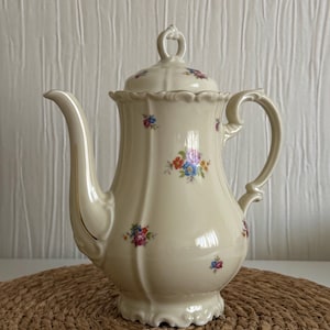 May include: A cream-colored porcelain teapot featuring a scalloped rim, a curved handle, and a matching lid with a finial. The body is decorated with small, scattered floral bouquets in shades of pink, blue, yellow, and green, with gold trim.