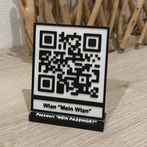 Könnte beinhalten: Ein schwarz-weißes QR-Code-Schild mit dem Text "Wlan Mein Wlan" und "Passwort MEIN PASSWORT". Das rechteckige Schild steht auf einem kleinen Sockel. Der Hintergrund ist eine hellfarbene Holzoberfläche.