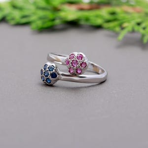 Op de afbeelding: Een zilveren ring met twee clusters edelstenen. De ene cluster is blauw en de andere is roze.