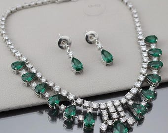 Puede incluir: Un conjunto de collar y pendientes plateados. El collar presenta piedras verdes esmeralda en forma de lágrima, intercaladas con pequeños cristales transparentes. Los pendientes tienen un diseño similar, con una sola piedra verde esmeralda.