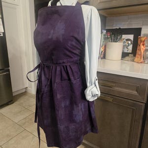 May include: A dark purple apron with two front pockets and a waist tie. The apron is displayed on a mannequin, showcasing its design and fit. The apron is made of a textured fabric.