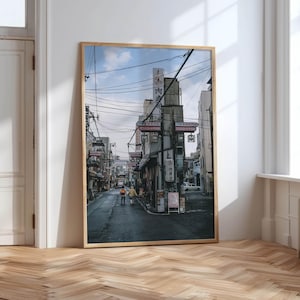 May include: Framed print of a Japanese street scene. The image features a narrow street lined with buildings, overhead power lines, and signage in Japanese. Two people walk down the street. The sky is overcast. The frame is a light wood color.