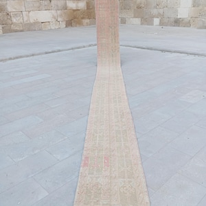 Könnte beinhalten: Ein langer, schmaler Teppich mit rechteckiger Form und verblichenem, vintage Aussehen. Der Teppich hat ein sich wiederholendes geometrisches Muster in Beige-, Rosa- und Rottönen. Der Teppich liegt auf einer grauen Steinoberfläche, die seine Länge und sein Design hervorhebt.