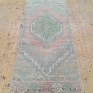 May include: A rectangular runner rug featuring a central diamond motif in shades of green, pink, and gray. The rug displays geometric patterns, including small diamond shapes and border designs, with a faded, weathered appearance throughout.