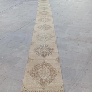 May include: A long, rectangular beige runner rug with an intricate, repeating geometric pattern in shades of brown. The rug is placed outdoors on a stone surface, showcasing its design and texture. Ideal for hallways or entryways.