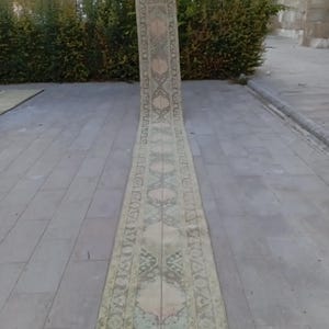Peut inclure: Un long tapis rectangulaire d'aspect délavé et vintage. Le tapis présente un motif central de motifs floraux et géométriques répétés dans des tons beige, vert et rose. Le tapis est exposé à l'extérieur sur une surface en pierre.