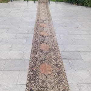 May include: An extra-long, narrow runner rug featuring a repeating pattern of large, faded peach-coloured medallions centred within dark charcoal and beige geometric borders. The rug has a weathered, distressed texture and short white fringe ends.