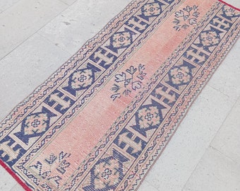 Puede incluir: Una alfombra larga y estrecha con fondo rosa y un patrón geométrico azul y blanco. La alfombra tiene flecos en ambos extremos.