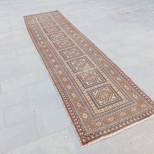 Puede incluir: Una alfombra larga y rectangular con un patrón geométrico marrón y crema. La alfombra tiene un borde de flecos y está colocada sobre un suelo de baldosas grises.