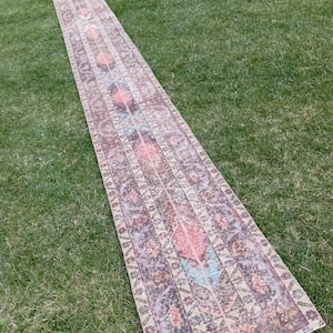 Könnte beinhalten: Ein langer, schmaler Vintage-Teppich mit verblasstem, abgenutztem Aussehen. Der Teppich hat ein zentrales Design mit floralen und geometrischen Mustern in gedämpften Rosa-, Blau- und Brauntönen. Der Teppich liegt auf einer grünen, grasbewachsenen Oberfläche.