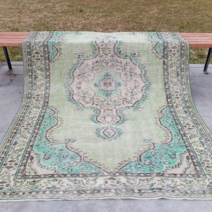 Könnte beinhalten: Ein großer, rechteckiger Teppich mit einem aufwendigen, verblassten Blumenmuster. Der Teppich hat ein hellgrünes Mittelfeld mit türkisfarbenen und braunen Akzenten. Der Rand ist dunkelgrün mit einem sich wiederholenden Blumenmuster. Der Teppich liegt auf einer Holzbank.